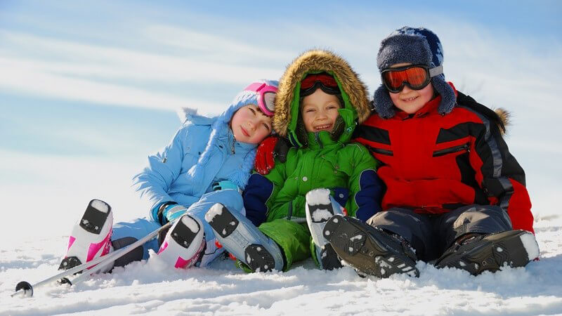 Drei kleine Kinder sitzen in Schneeanzügen und mit Skischuhen im Schnee