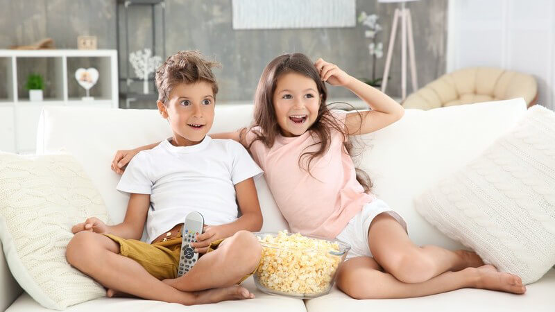 Bruder und Schwester sitzen gemütlich mit einer großen Schüssel Popcorn auf der weißen Couch und gucken Fernsehen