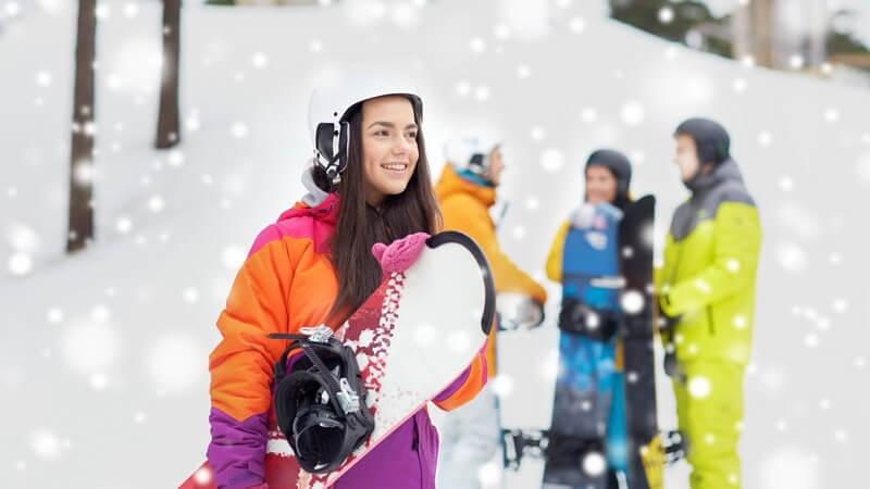 Junge Frau in Skibekleidung mit weißem Helm und Snowboard unter dem Arm, im Hintergrund stehen drei Freunde
