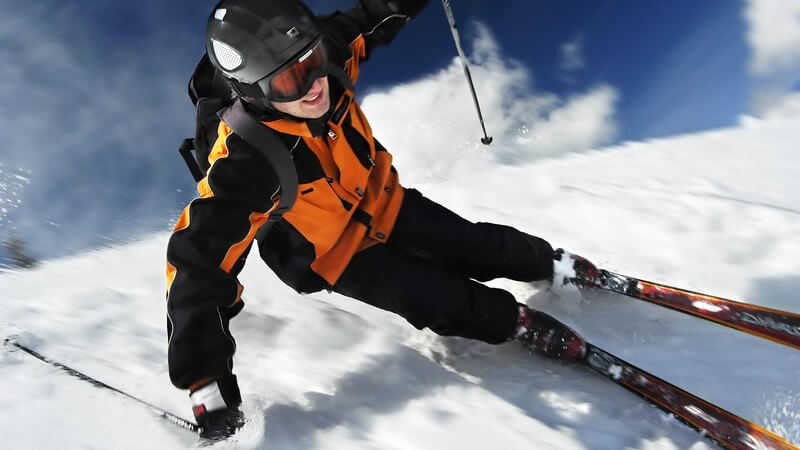 Skifahrer mit Helm und Skibrille carvt unter blauem Himmel eine Piste hinunter