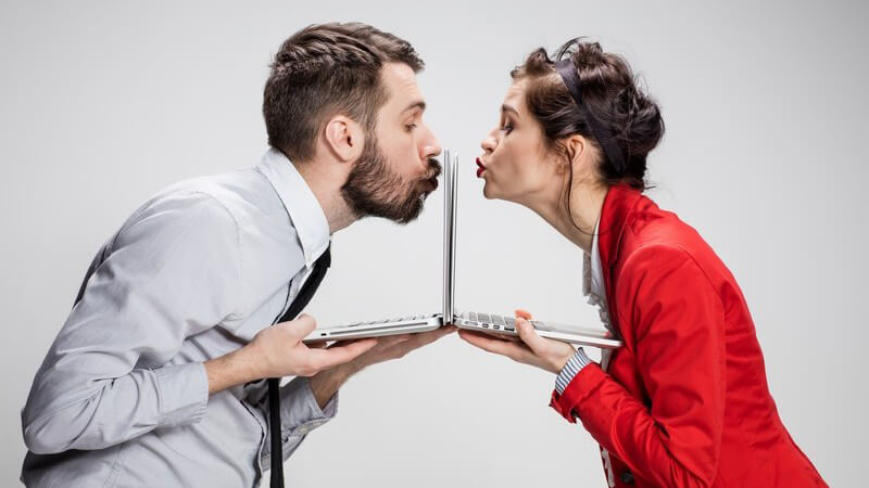 Mann und Frau in Businesskleidung stehen sich mit ihren Laptops direkt gegenüber und küssen jeweils ihren Bildschirm