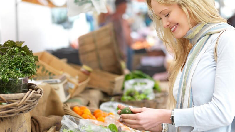 Junge, blonde Frau auf Gemüsemarkt hält Avocado in den Händen
