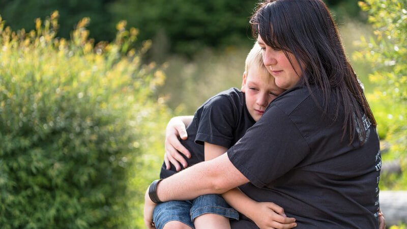 Weinender Junge sitzt auf dem Schoß der Mutter, sie umarmt und tröstet ihn