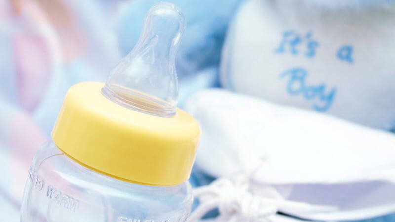 Babyfläschchen mit gelbem Rand, im Hintergrund Teddy mit Aufschrift "It's a Boy"
