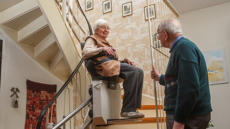 Senioren-Paar im Treppenhaus seines Zuhauses, Frau sitzt auf dem Treppenlift