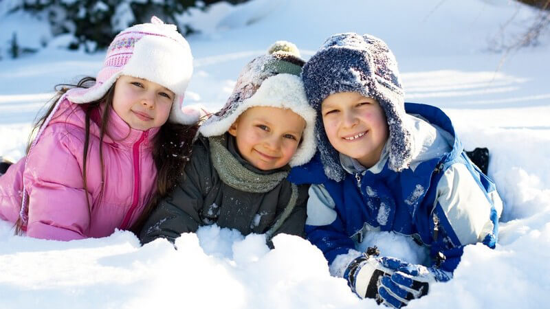 Drei kleine Kinder sitzen in Schneeangzügen im Schnee und lächeln in Kamera