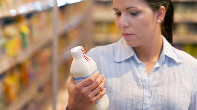 Frau im Supermarkt steht mit Einkaufswagen vor Regal und schaut sich Flasche an