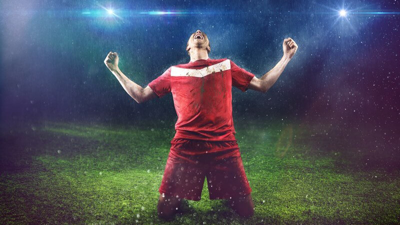 Fußballspieler in rotem Trikot kniet im Regen und unter Flutlicht auf dem Rasen und jubelt