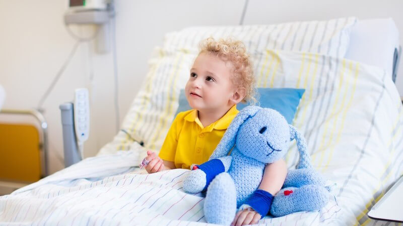 Kleiner Junge in gelbem Poloshirt sitzt mit seinem blauen Kuscheltier im Krankenhausbett