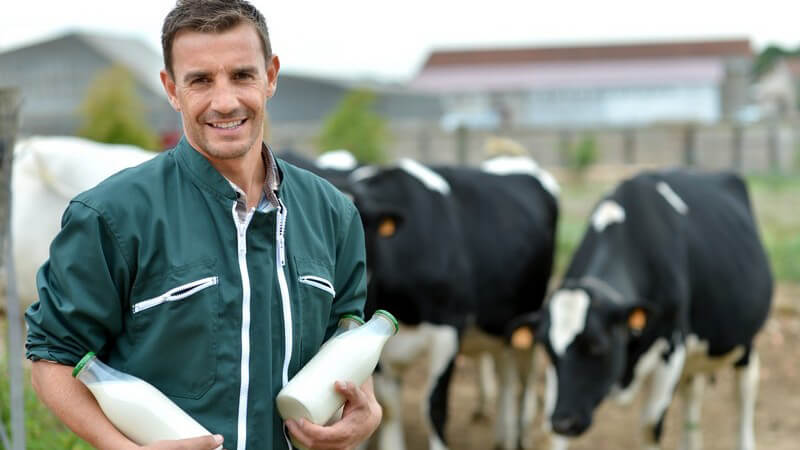 Landwirt in grünem Overall steht mit Milchflaschen in den Händen vor seinen Kühen