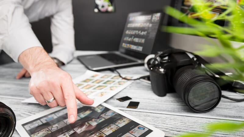 Fotograf zeigt auf Fotos auf einem Tablet, Laptop und Kamera liegen auf dem Schreibtisch