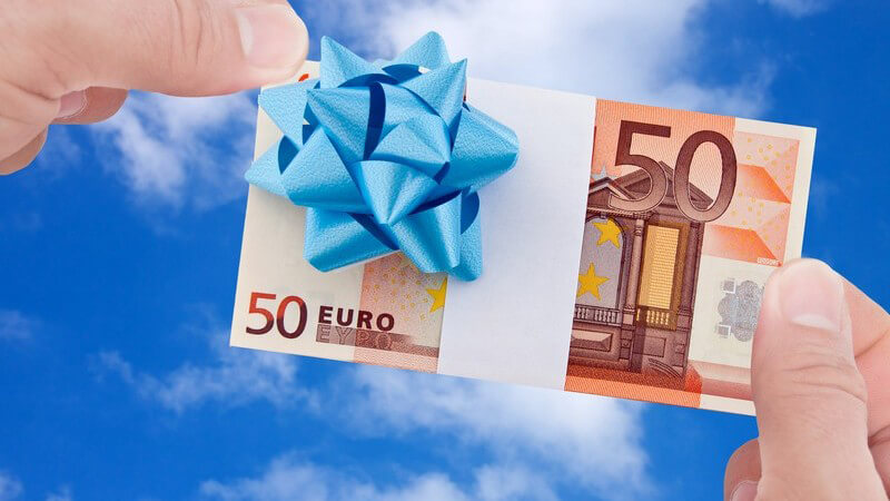 Zwei Hände halten einen 50-Euro-Geldschein mit weißem Band und blauem Schleifchen vor einen blauen Himmel