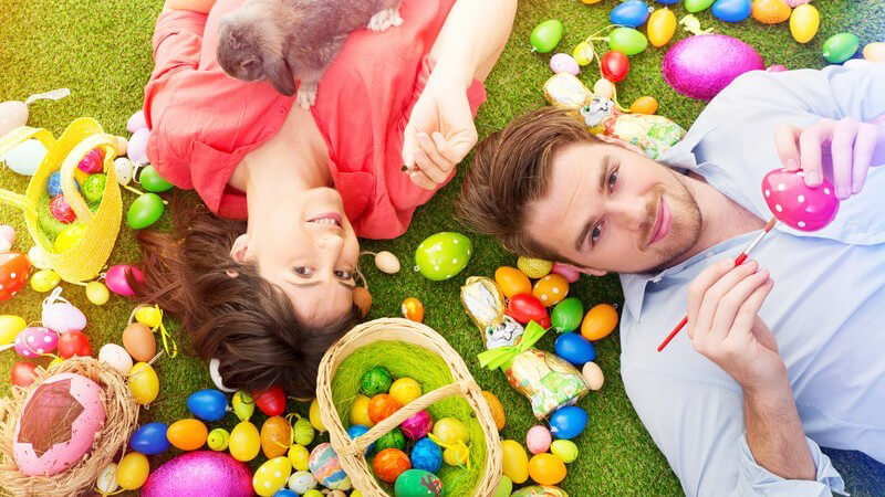 Paar liegt mit einem Kaninchen auf Kunstrasen, umgeben von vielen bunten Ostereiern