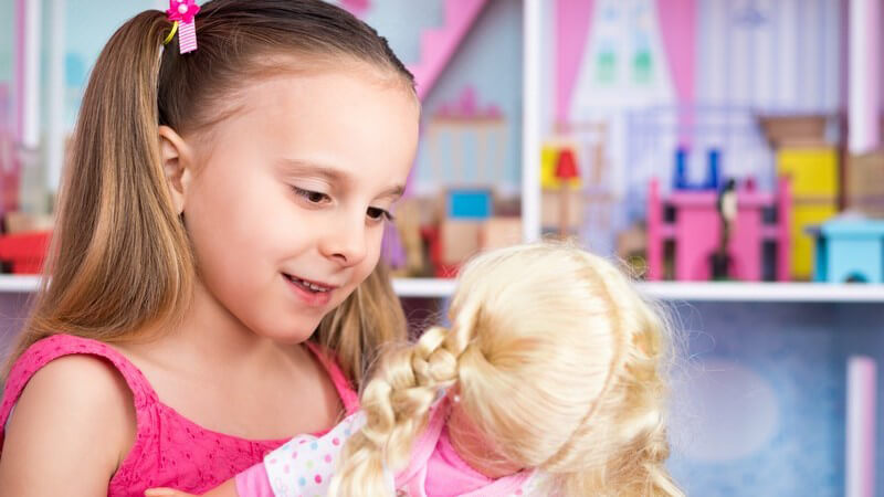 Mädchen mit Zöpfen spielt mit ihrer blonden Puppe, im Hintergrund steht ein Puppenhaus