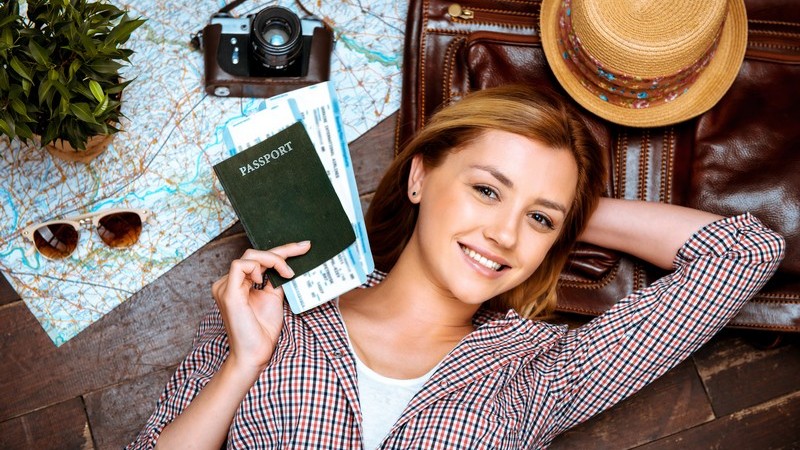 Jugendliche liegt neben Strohhut, Karte, Kamera und Reisetasche auf dem Boden und hält Reisepass und Flugtickets
