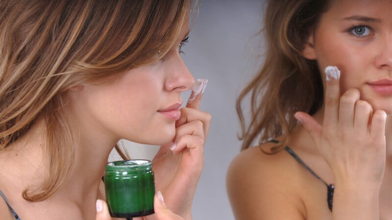 Junge Frau vor Spiegel trägt Gesichtscreme aus grünem Döschen auf