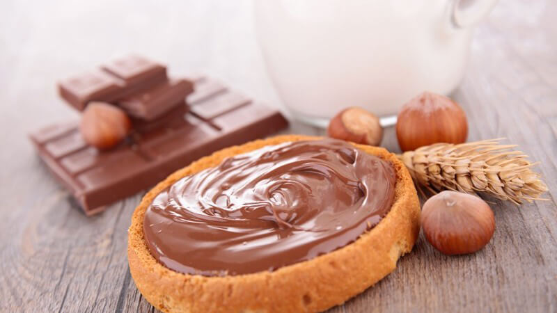 Brotaufstrich: Schokoladencreme oder Nougatcreme auf Brotscheibe, dahinter Schokolade und Haselnüsse