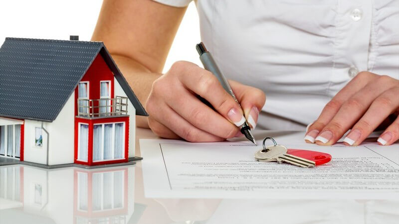 Frau unterschreibt einen Kauf- oder Mietvertrag für ein Haus, auf dem Tisch liegen ein Schlüssel und ein Modellhaus