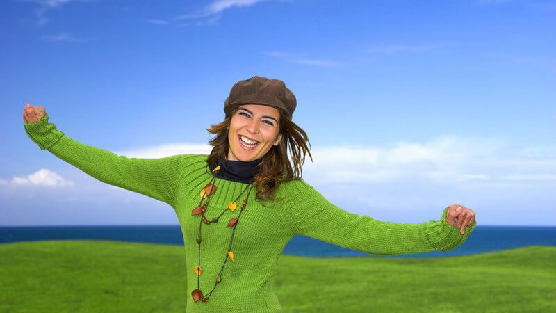 Junge fröhliche Frau mit Mütze, ausgestreckte Arme, im Hintergrund Wiesenlandschaft