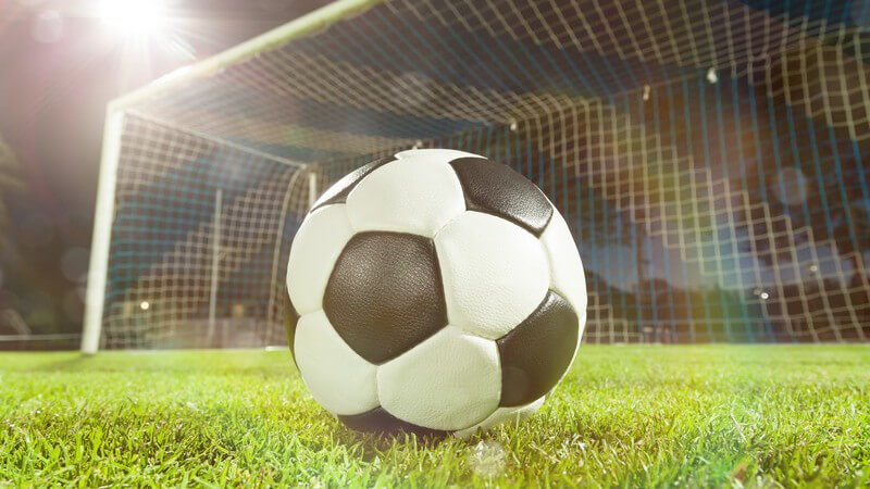 Ein Fußball liegt vor dem Tor auf dem Rasen und wird von einem Flutlicht angeleuchtet