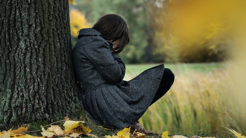 Herbstblues - Frau mit Winterdepression hockt traurig am Baumstamm auf Herbstwiese