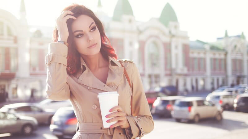 Junge Frau in beigem Mantel steht in der Morgensonne an einem Parkplatz und hält einen Kaffee in der Hand