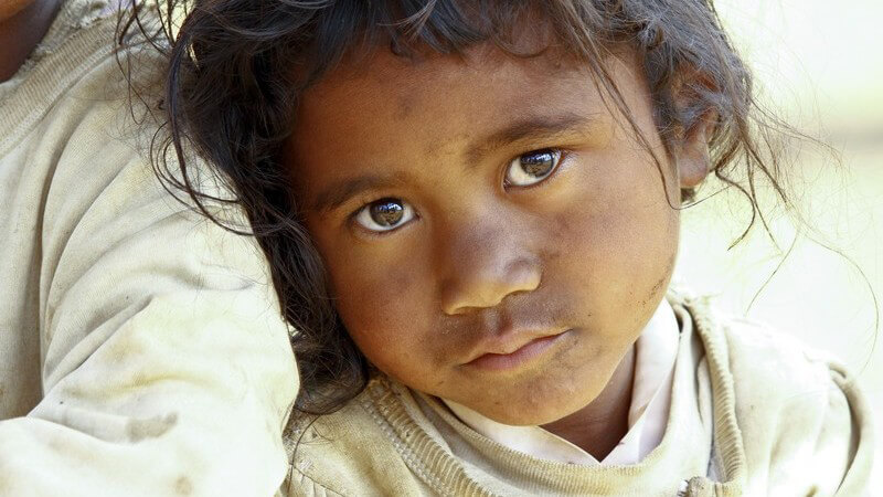 Kleines, dunkelhaariges Mädchen in schmutziger Kleidung lehnt traurig an einem anderen Kind