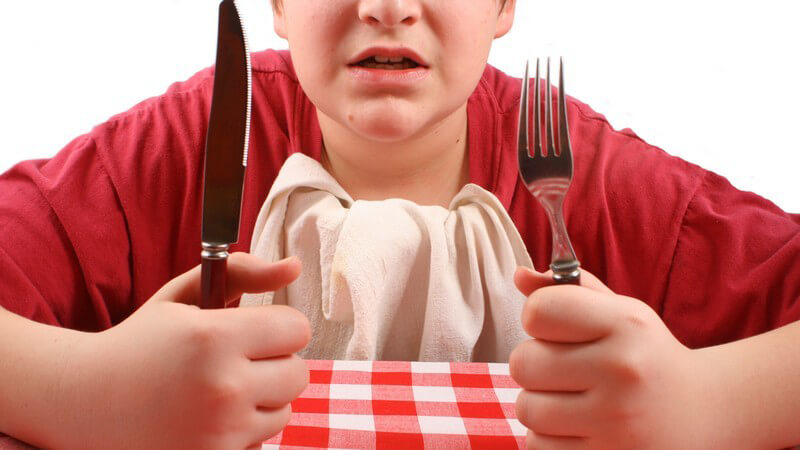 Junge sitzt mit Messer und Gabel am Tisch und wartet ungeduldig auf sein Essen