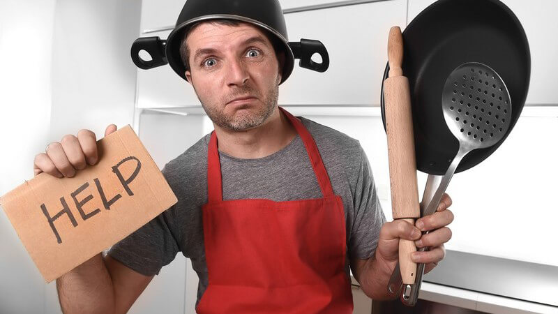 Mann steht mit roter Schürze und Kochtopf auf dem Kopf in der Küche und hält ein Schild mit der Aufschrift "Help" hoch