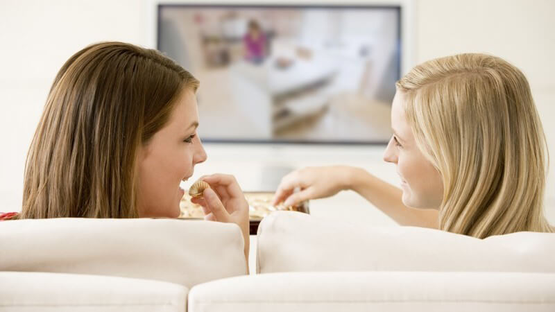 Rückenansicht zwei Freundinnen auf Couch vor Fernsehen essen Pralinen