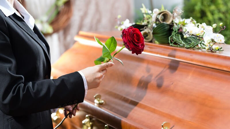 Frau in Schwarz legt bei einer Beerdigung eine rote Rose auf einen Holzsarg