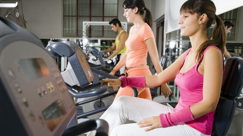 Brünette Frau im pinken Outfit sitzt im Fitnessstudio, zwei weiter Personen trainieren dahinter