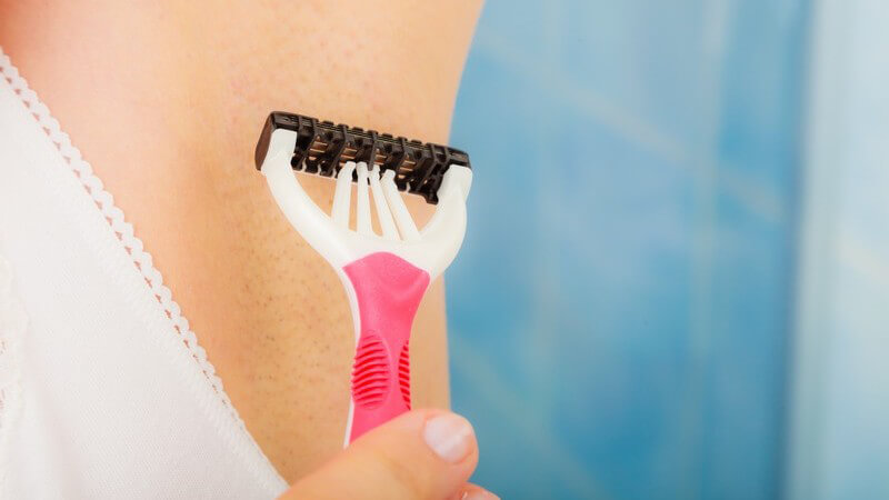 Frau rasiert sich mit weiß-pinkem Nassrasierer die Achselhöhle im Badezimmer