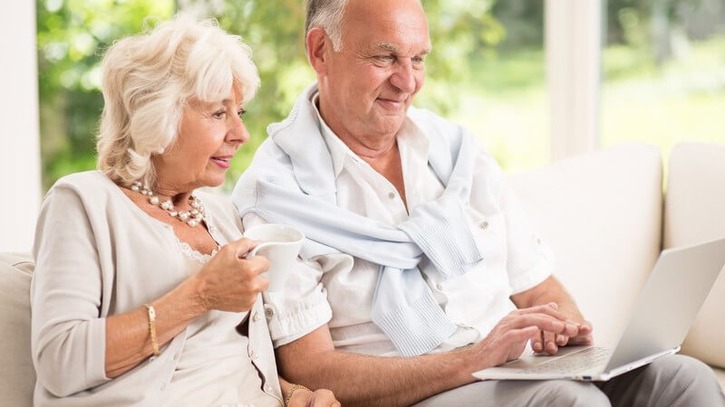 Altes Ehepaar sitzt auf der Couch, sie hält eine Tasse, er einen Laptop
