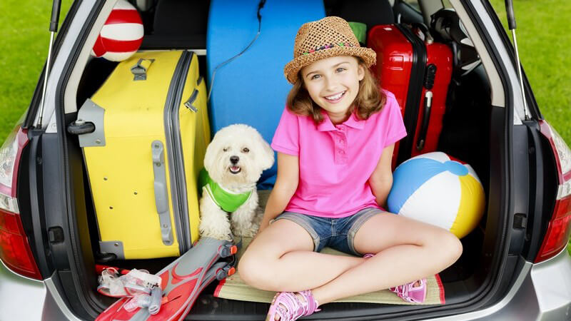 Mädchen mit Strohhut und Hund sitzen in einem vollgepackten Kofferraum