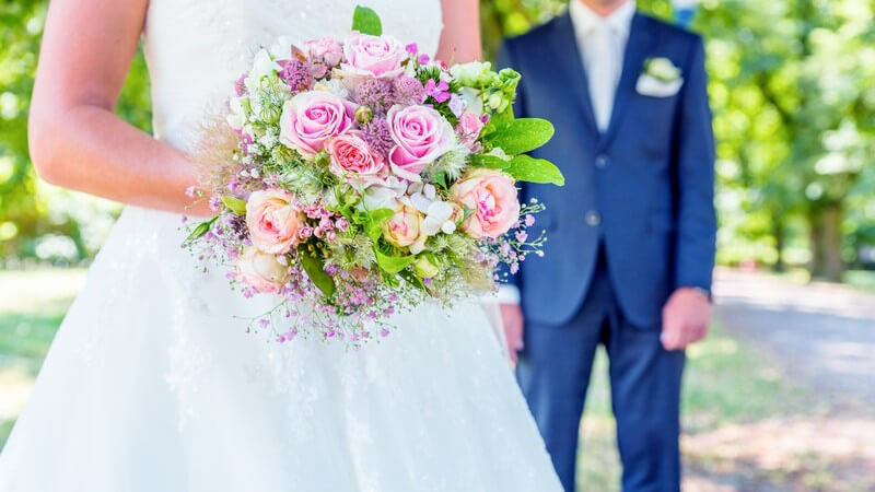 Weiße Braut mit Brautstrauß im Grünen, im Hintergrund der Bräutigam in blauem Anzug