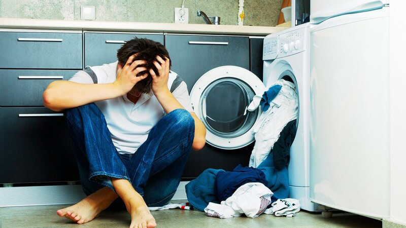 Verzweifelter junger Mann sitzt vor Waschmaschine in Wohnung, aus der Wäsche kommt, hält sich Kopf, barfuß