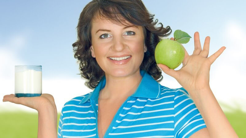 Dunkelhaarige, lächelnde Frau hält grünen Apfel und ein Glas Milch