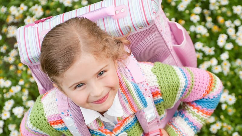 Kleines Mädchen mit Schulrucksack steht in Blumenwiese und schaut nach oben in Kamera