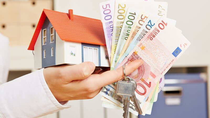 Hand im Büro hält ein Hausmodell, Euro-Geldscheine und Schlüssel