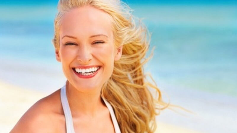 Junge, lächelnde blonde Frau mit weißem Bikini am Strand vor azurblauem Meer