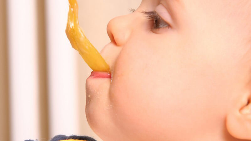 Kleinkind wird gefüttert- Nahaufnahme vom Kopf mit orangenem Plastiklöffel im Mund