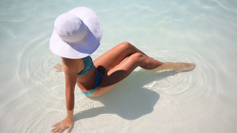 Frau im Bikini mit Sonnenhut sitzt in flachem Wasser