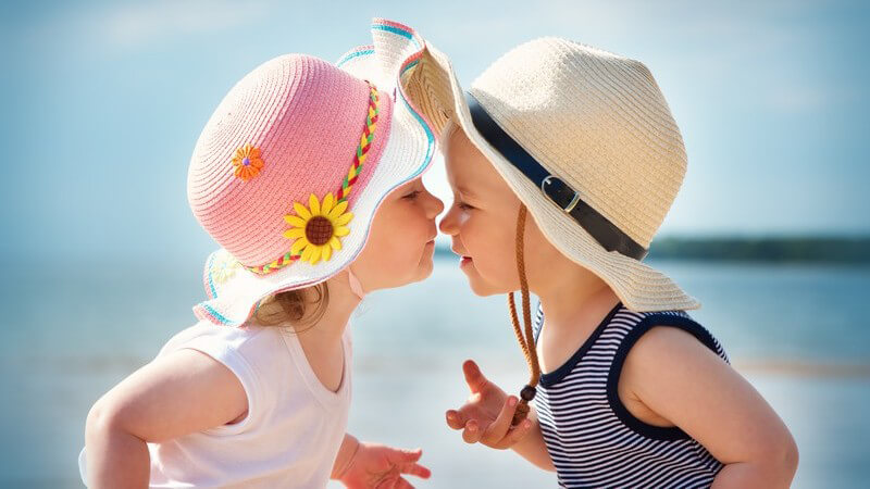 Kleines Mädchen und kleiner Junge, beide mit Sonnenhut, küssen sich am Strand