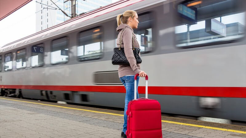 Frau mit Handtasche rotem Trolley wartet am Bahnsteig auf ihren Zug