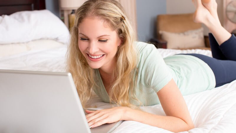 Blonde Frau mit Haarspange liegt lächelnd auf dem Bett vor einem Laptop
