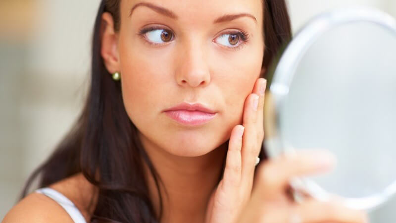 Junge Frau schaut in Kosmetikspiegel, streicht sich über Wange