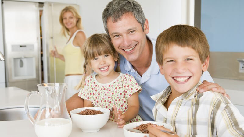 Vater mit zwei kleinen Kindern beim Frühstückstisch, im Hintergrund Mutter an Kühlschranktür