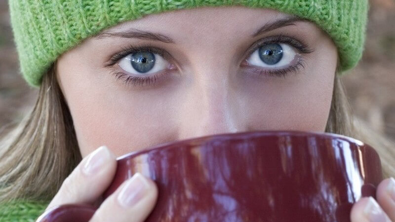 Nahaufnahme von junger Frau mit grüner Wollmütze und blauen Augen, die aus bordeauxfarbener Kaffeetasse trinkt