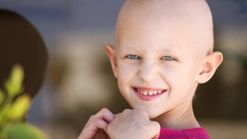 Krebskrankes Kind mit pinkem Oberteil und ausgefallenen Haaren (Glatze) blickt lächelnd in die Kamera